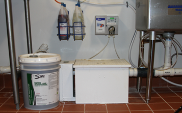 A gallon bucket of a State Chemical branded bucket is pictured next to a white rectangular box.  To the right of the box, there is the bottom of a sink.  The State branded box is hooked up to the sink through a clear tube.  The tube slowly dispenses product from the bucket to the sink drain.